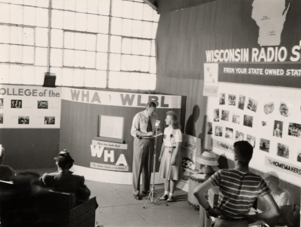 A man and woman stand at microphone in the midst of a WHA display at a state fair.