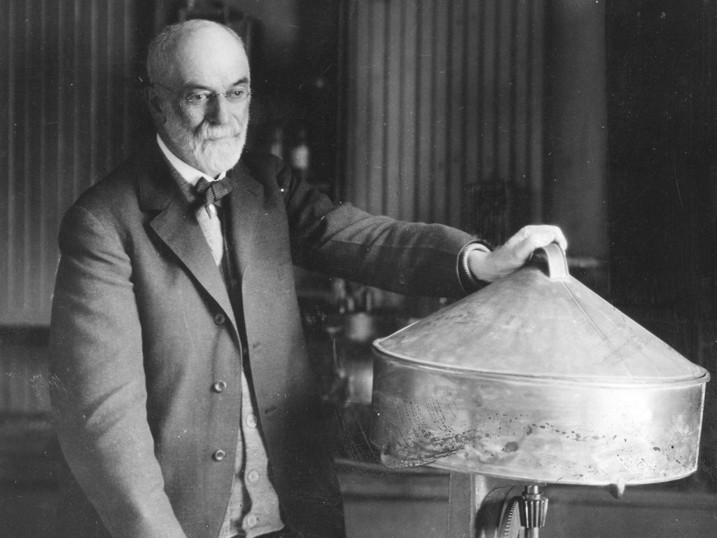 Black and white photo of an elderly, bearded man in a suit resting his hand on a large, metallic butter-churning device.