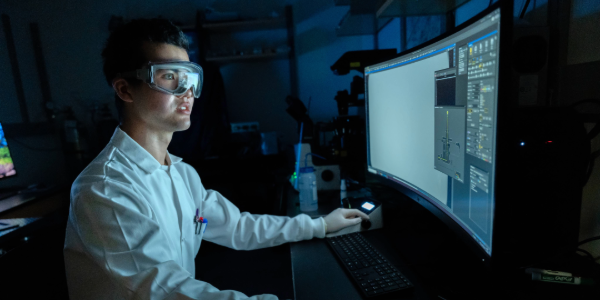 A student wearing a white lab coat and goggles is sitting in a dark lab at a computer looking at the monitor